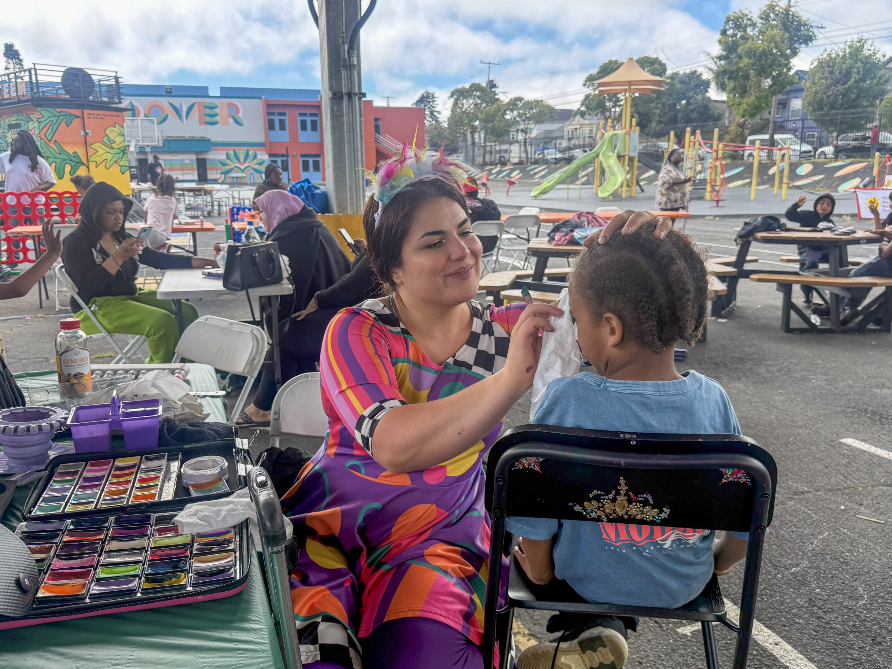 A woman wearing colorful clothing and a feathery headband does face paint on a child at Hoover Elementary School